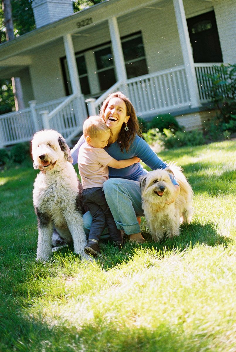 Laughing woman sitting on grass holding baby with two fluffy dogs beside her in front of a white house on sunny day.