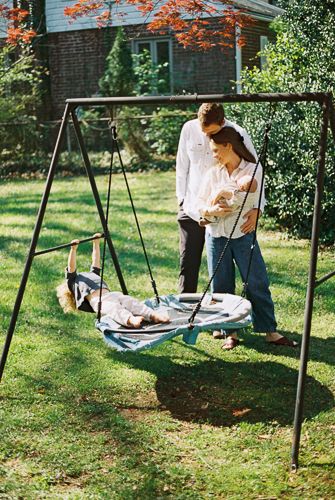 A couple stands together in a backyard holding a baby near a swing set on a sunny day.