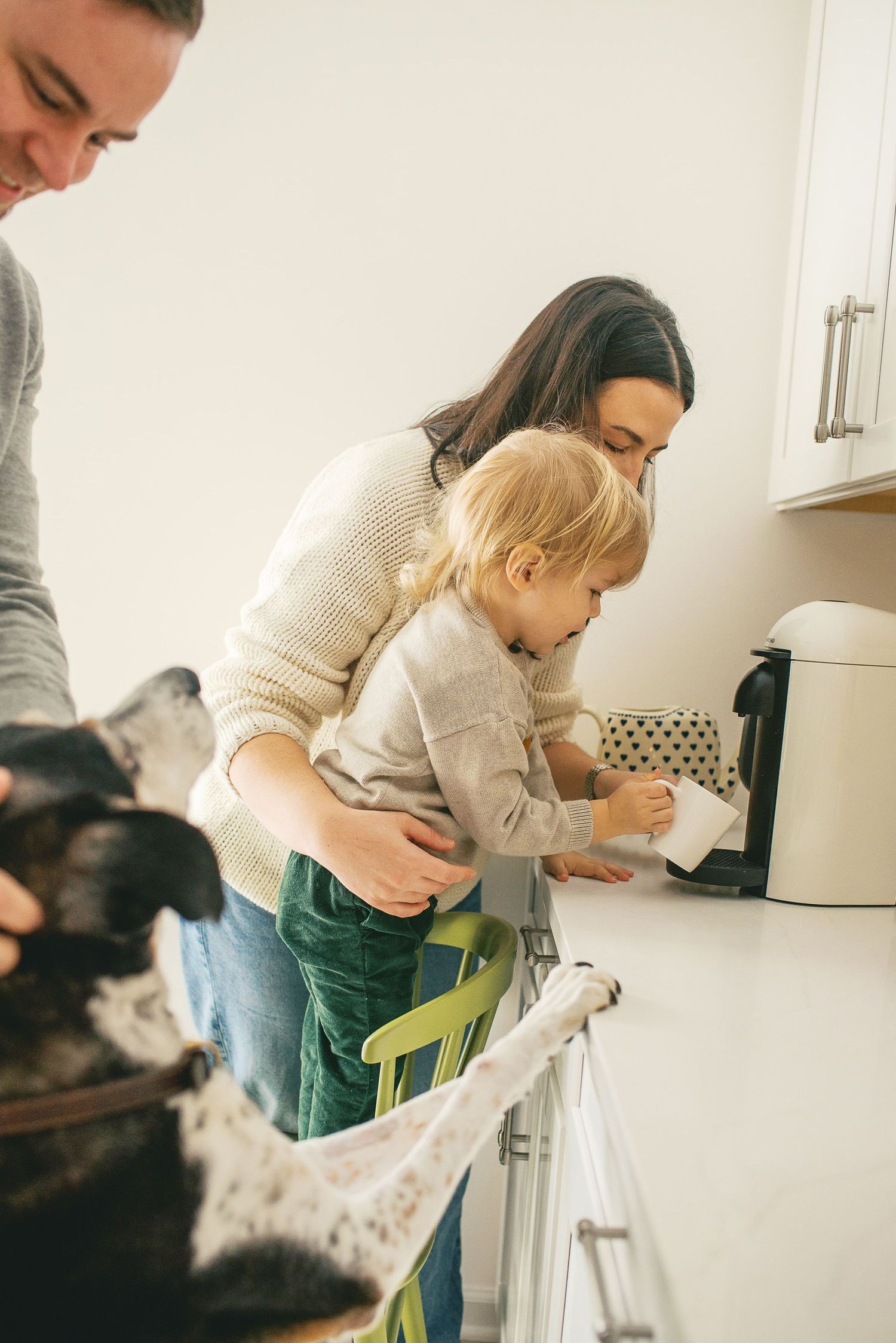 Family cooking together in bright kitchen with toddler standing on step stool while dog watches nearby.
