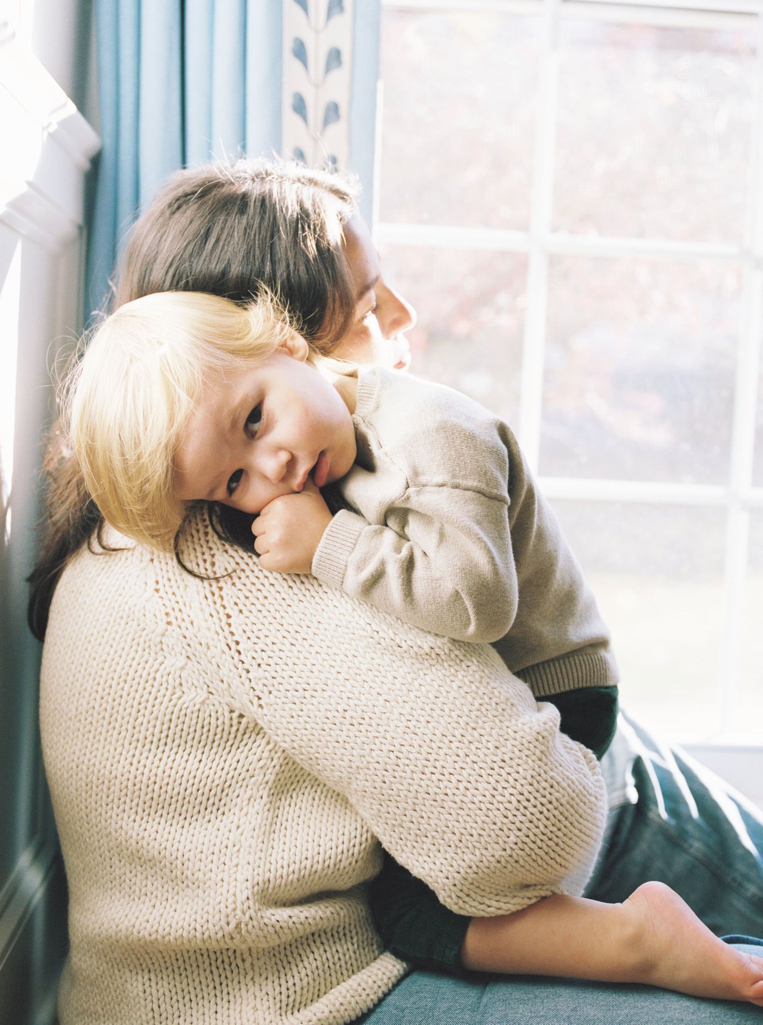 A tender moment by a window shows a gentle embrace between two people wearing cozy knit sweaters in soft natural light.