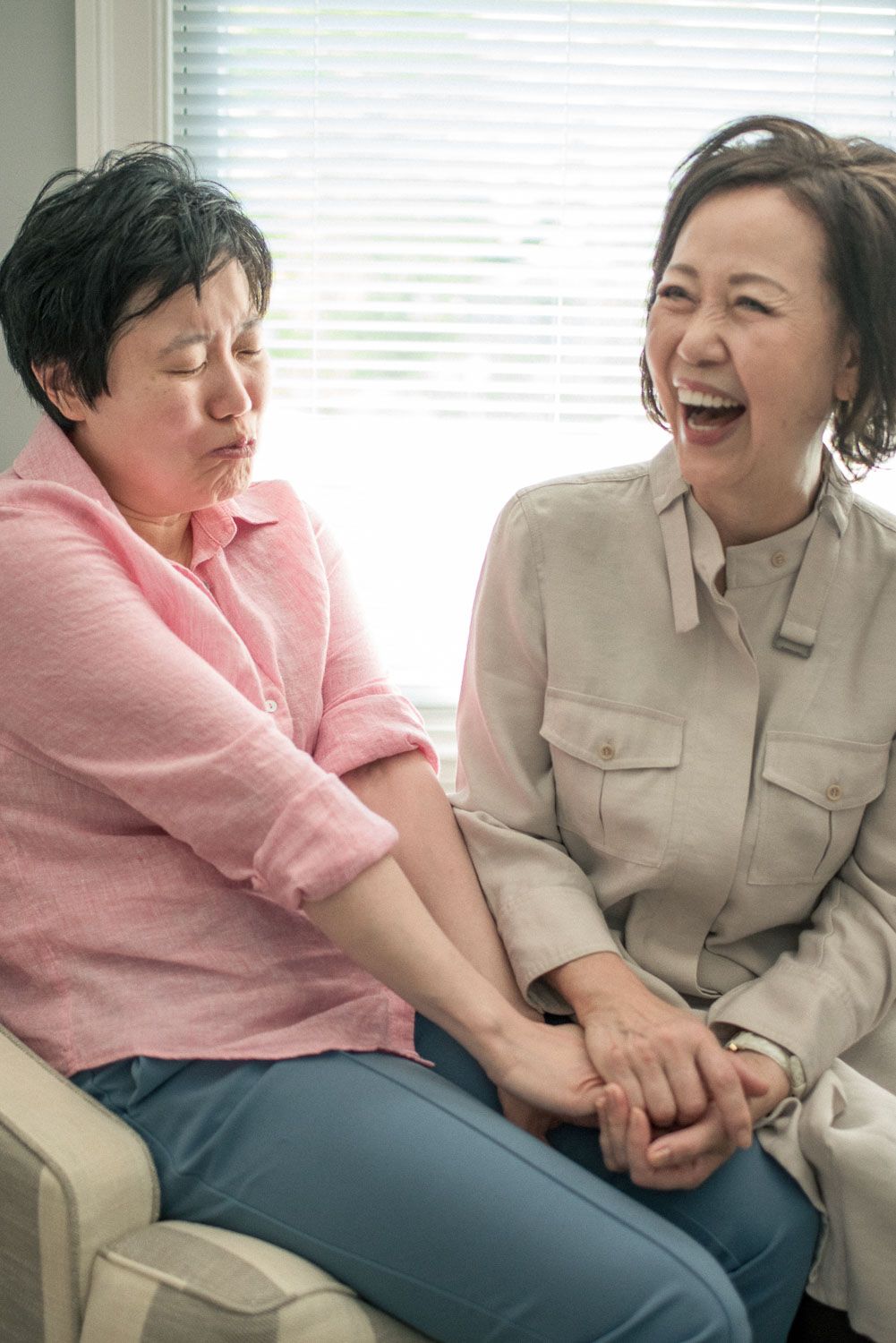 Two Asian women laughing and holding hands while sitting together on a couch near a window.