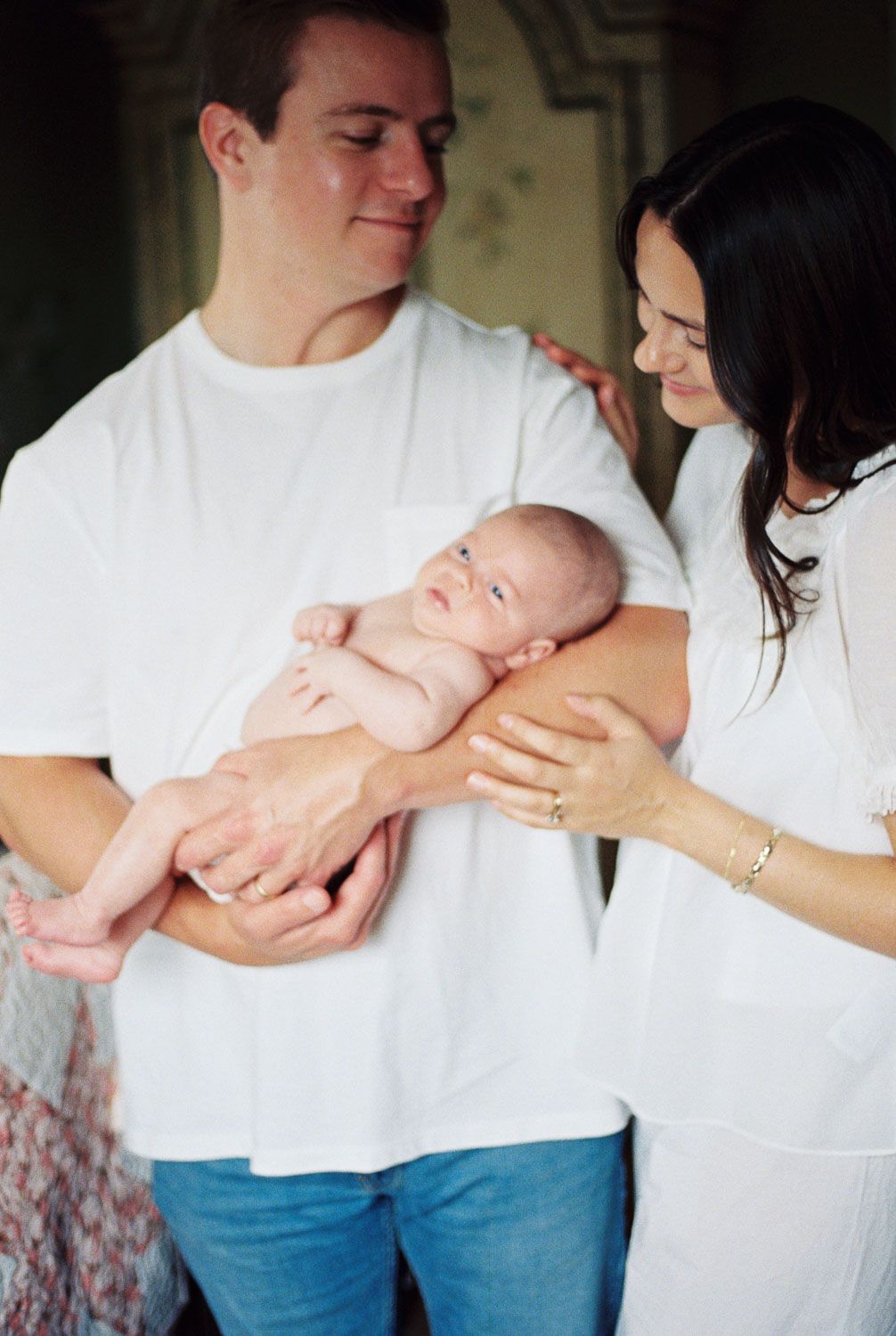 A young couple in white shirts tenderly holds their newborn baby, sharing a loving family moment together.