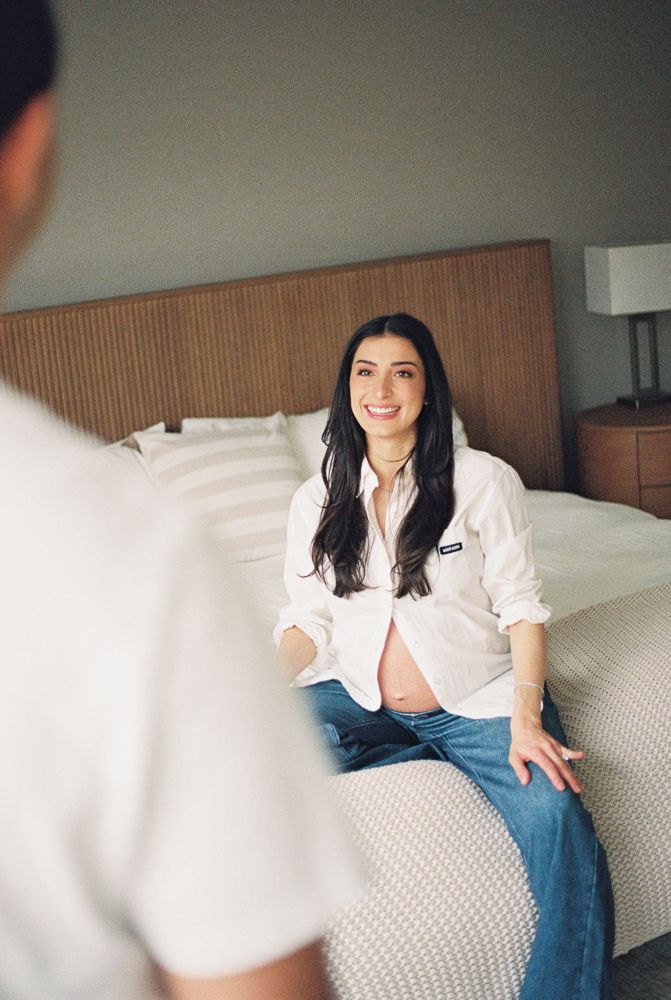 Person in white button-down shirt and jeans sits casually on white couch in modern bedroom setting.