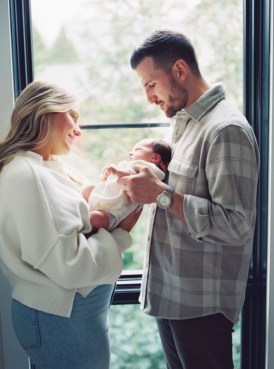 A couple shares a tender moment while holding their newborn baby near a bright window.