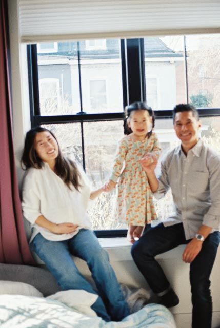 A happy family of three poses by a window, with parents holding their young daughter in a floral dress indoors.
