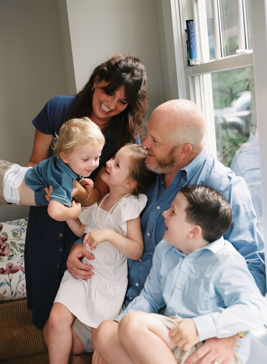 Family shares tender moments of laughter and affection while cuddling together on a light-colored sofa in a sunlit room.