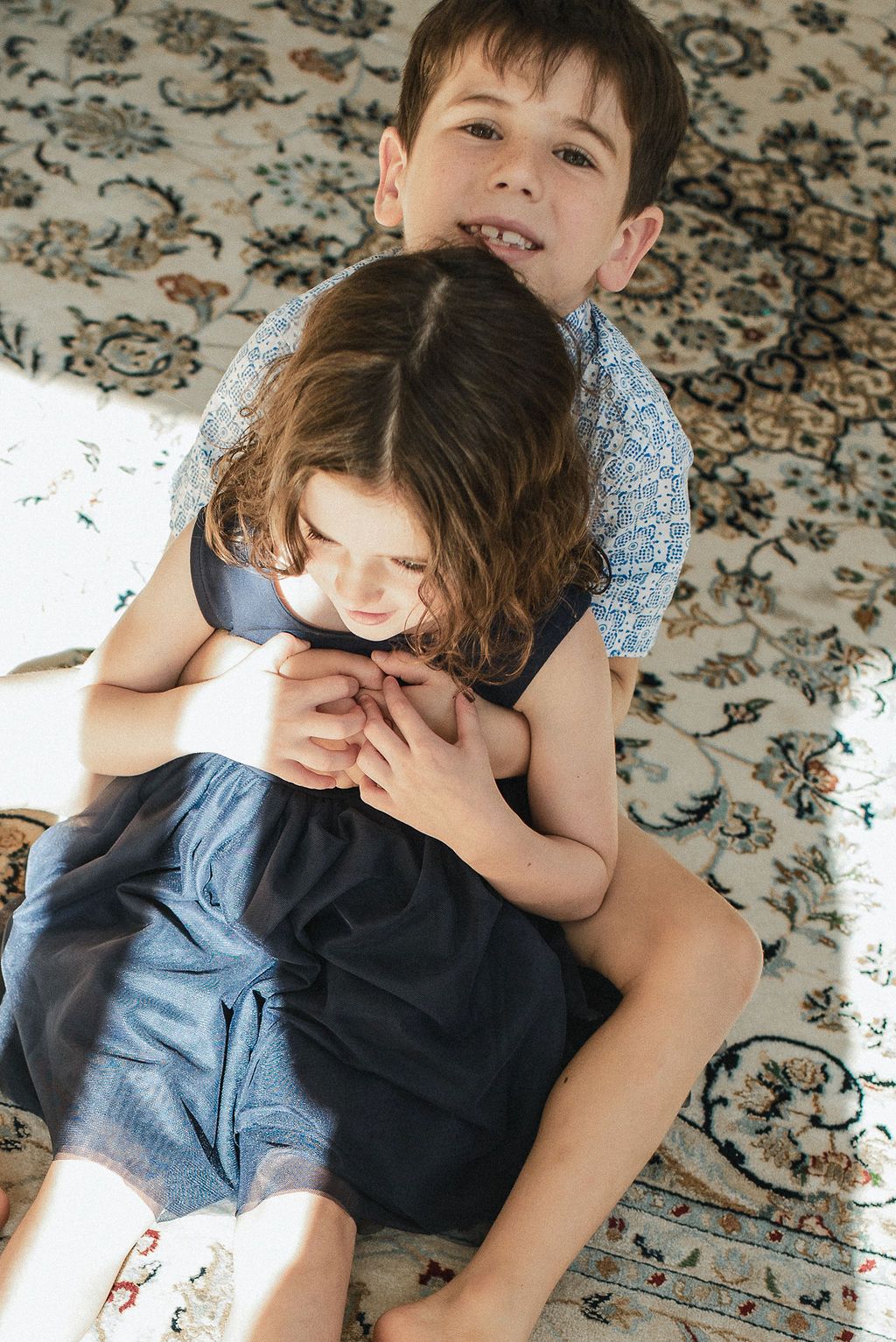 Two children lying on a patterned rug share a sweet embrace while playing together.