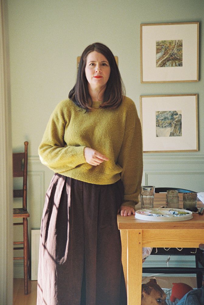Someone wearing a mustard yellow sweater and brown skirt stands in a room with framed artwork on the wall.