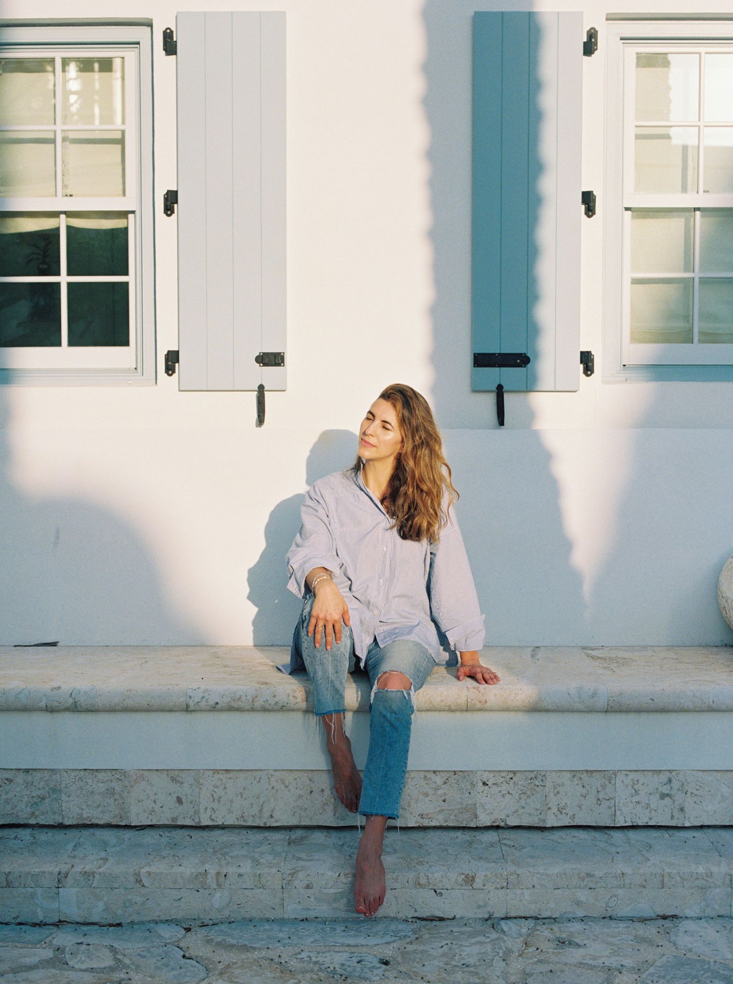 A person in jeans and a sweater poses against a white building wall with blue shutters in a series of creative shots.