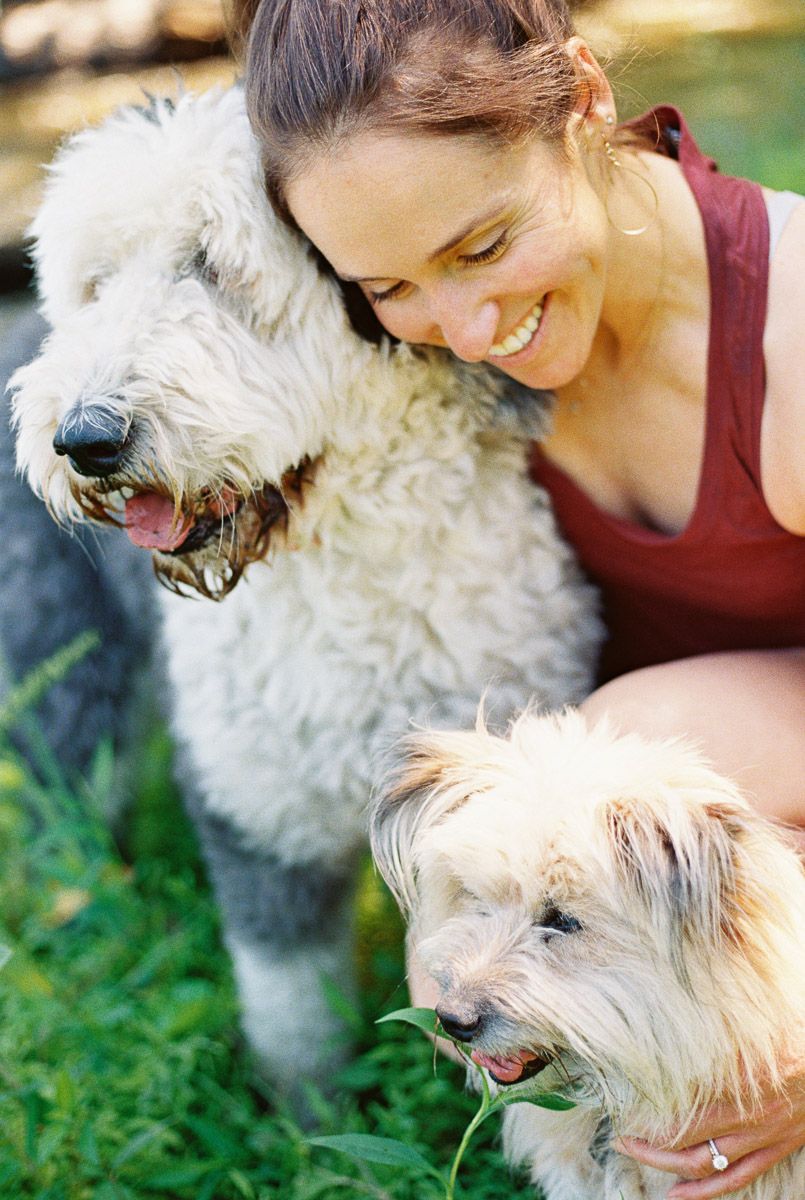 Smiling woman in red top hugging two fluffy white dogs on green grass outdoors.