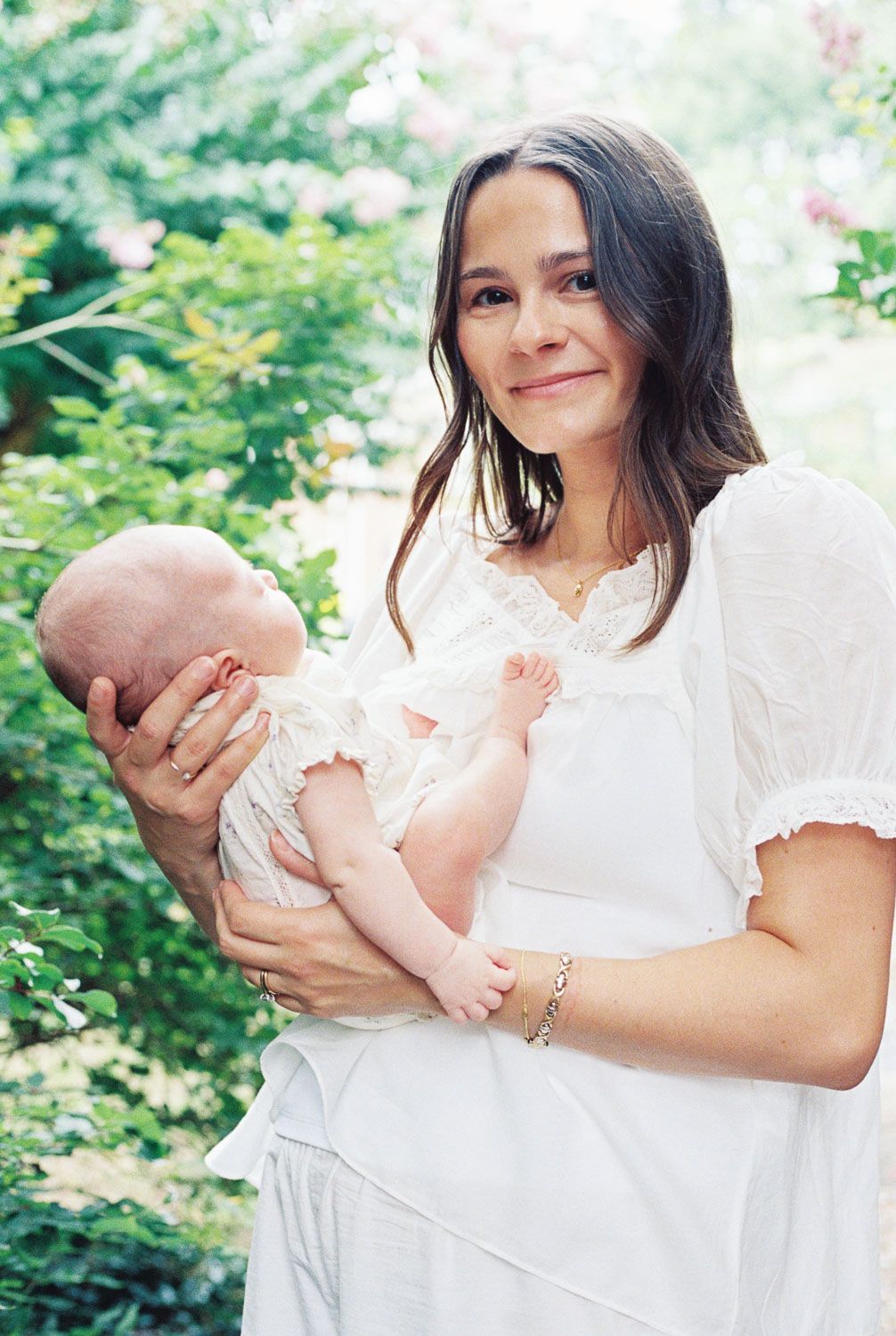 A smiling brunette woman in a white lace dress holds a sleeping newborn baby outdoors among lush green foliage.
