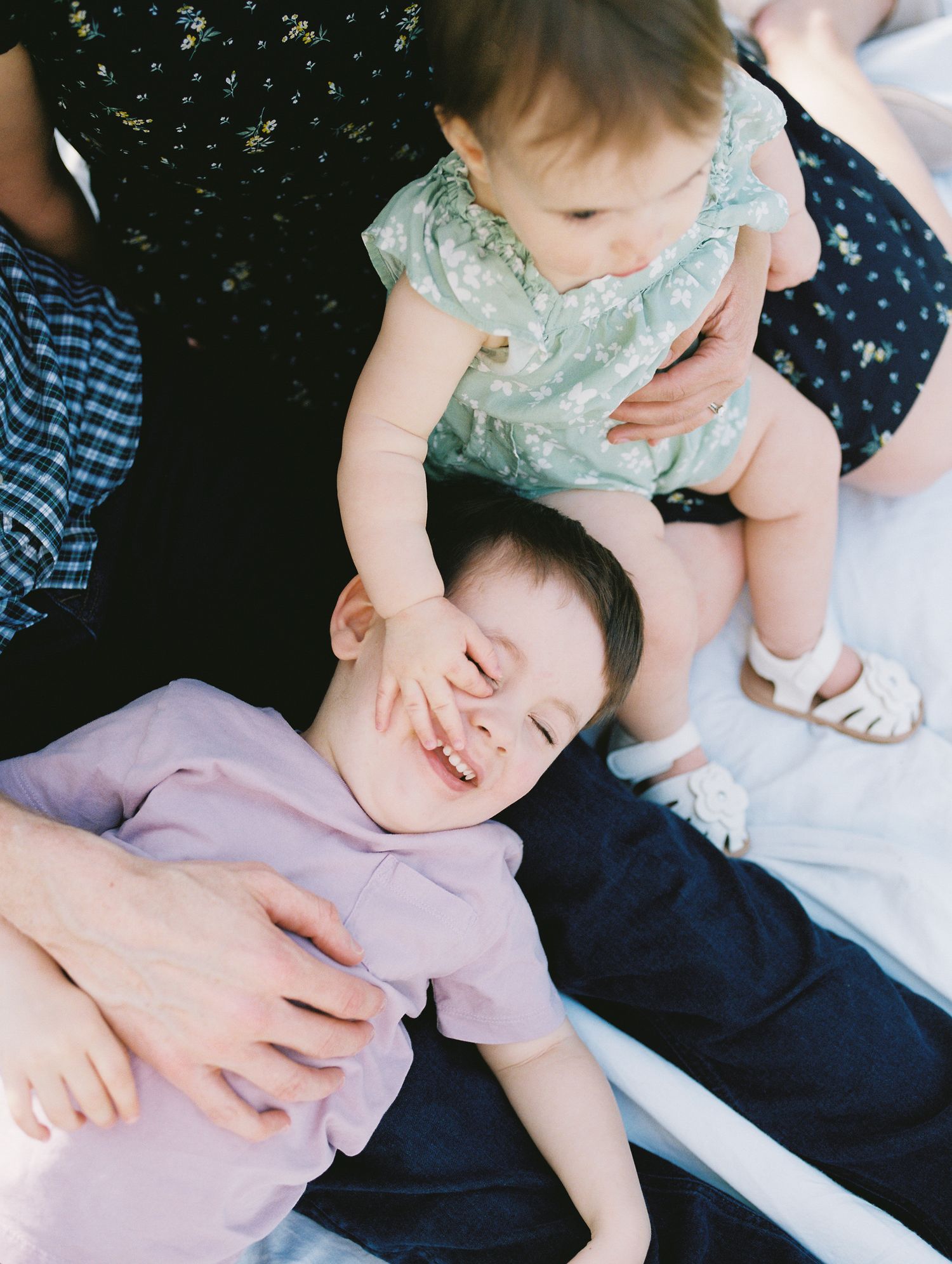 Baby in mint green outfit sits near someone resting on a bed in lavender shirt and navy blue pants.