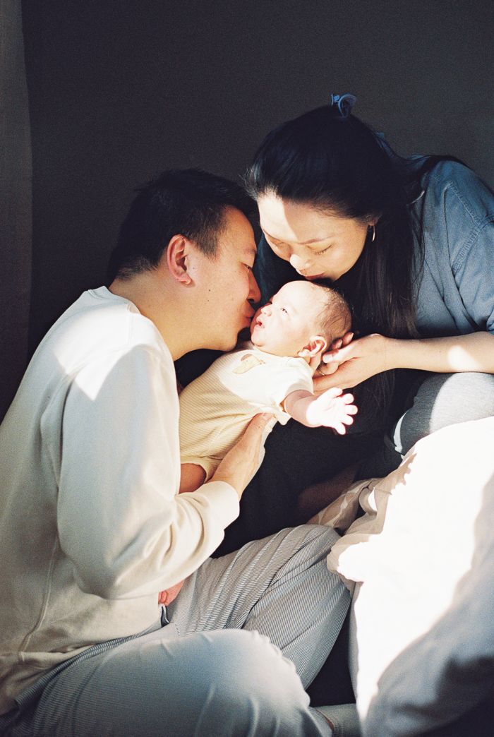 Two parents lovingly kiss their newborn baby in a warm, intimate family portrait captured on film.