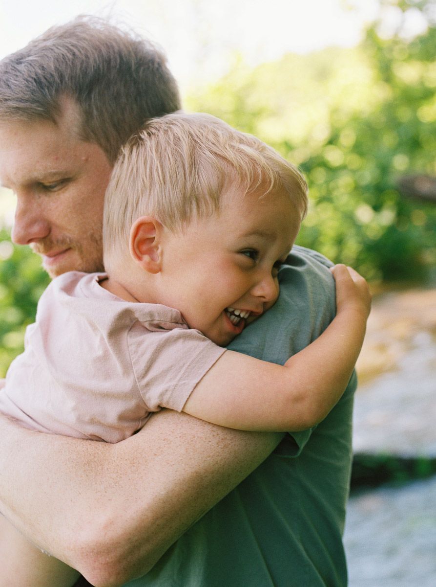 A smiling young boy hugs a man outdoors, surrounded by lush green foliage in a warm, candid moment.