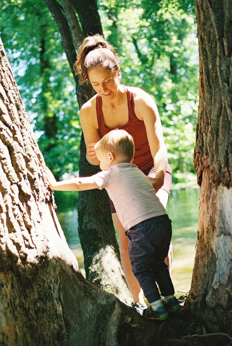 A mother helps her toddler climb a tree trunk near a river in a sunny park.