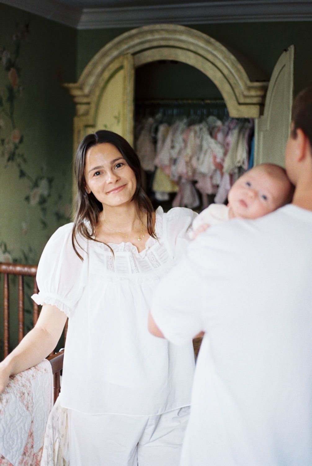 A woman in a white dress smiles warmly as another person holds a newborn baby in a vintage-style room.
