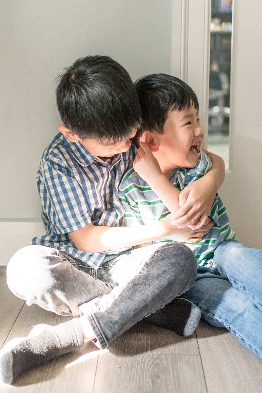 Two young Asian boys sitting on floor, older brother hugging younger sibling who is smiling.