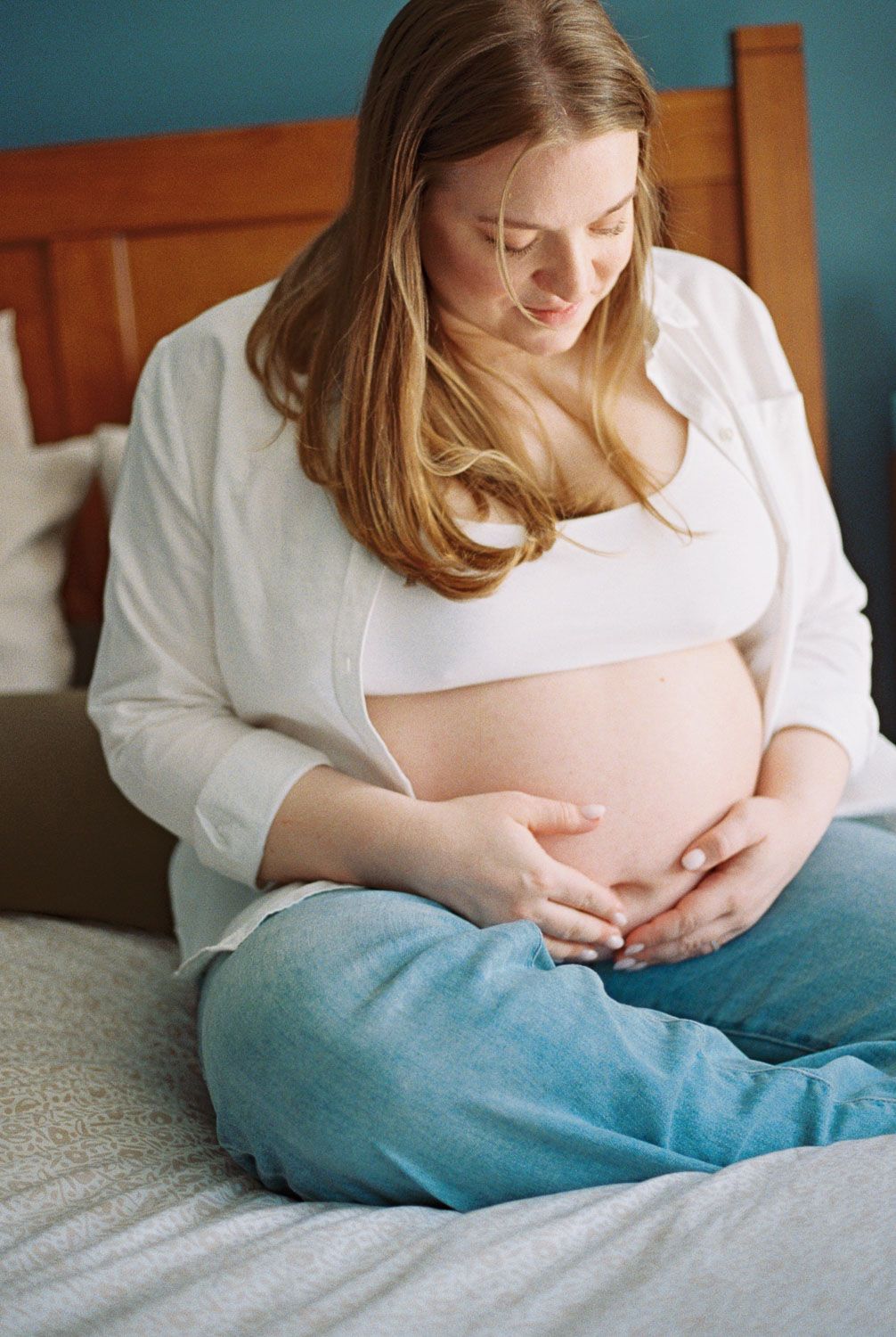 Pregnant woman sitting on bed cradling her bare baby bump, wearing white shirt and blue jeans.