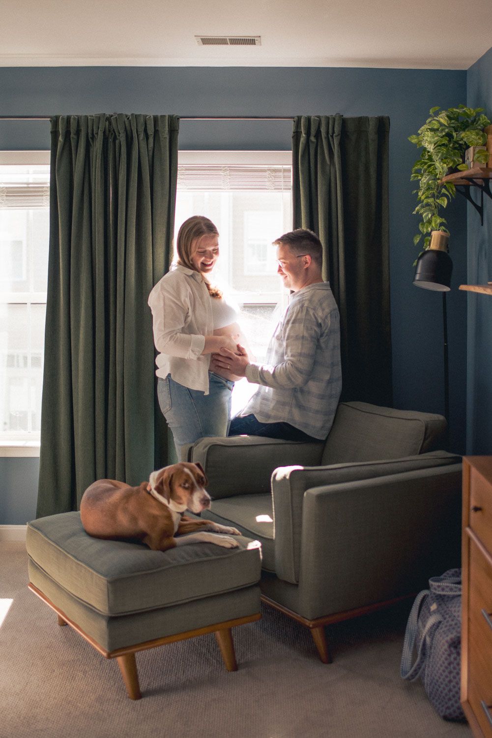 Happy couple touching pregnant belly in a cozy blue living room with a beagle dog resting on an ottoman.