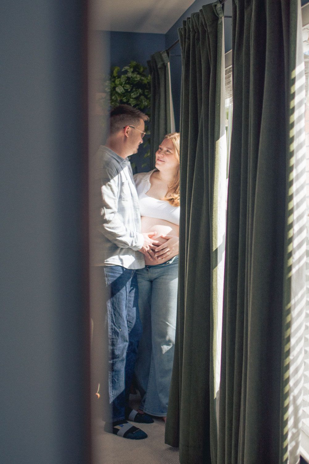 Couple sharing a tender moment by a sunlit window, with the woman's pregnant belly visible in a cozy room.