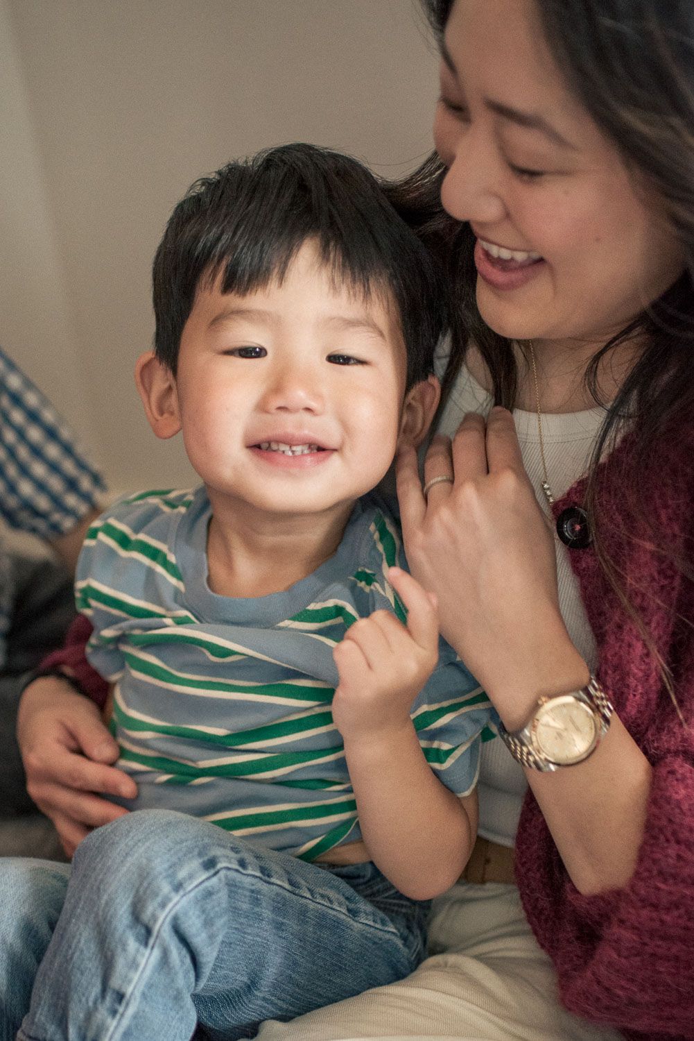 Happy Asian toddler boy in striped shirt smiling while sitting with his mother.