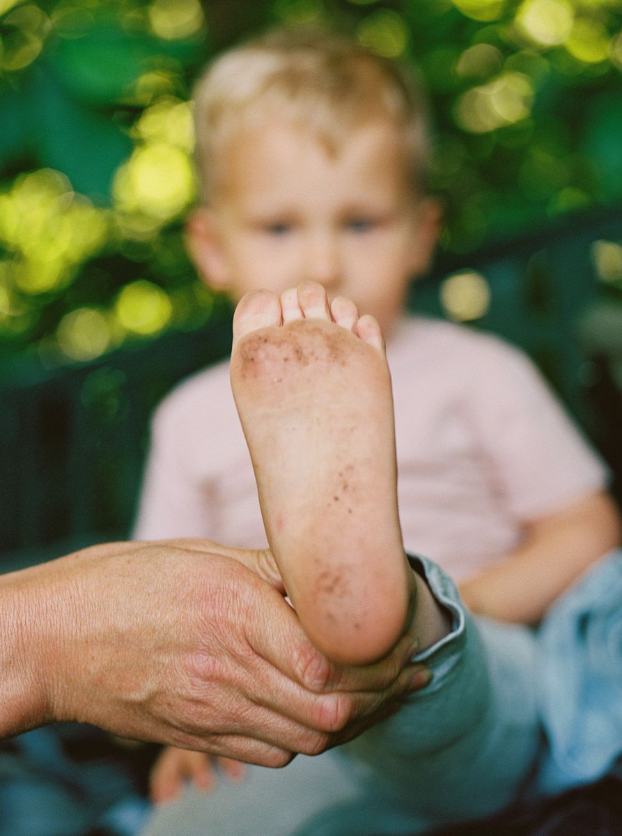Adult hands hold a young toddler's dirty bare foot outdoors with a blond child visible in background.