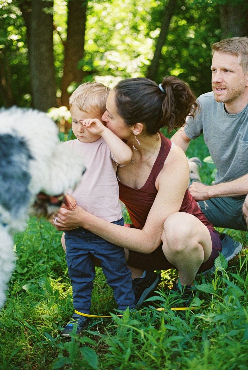 A mother kisses her toddler outdoors as father watches, with a fluffy dog nearby in a lush green forest setting.
