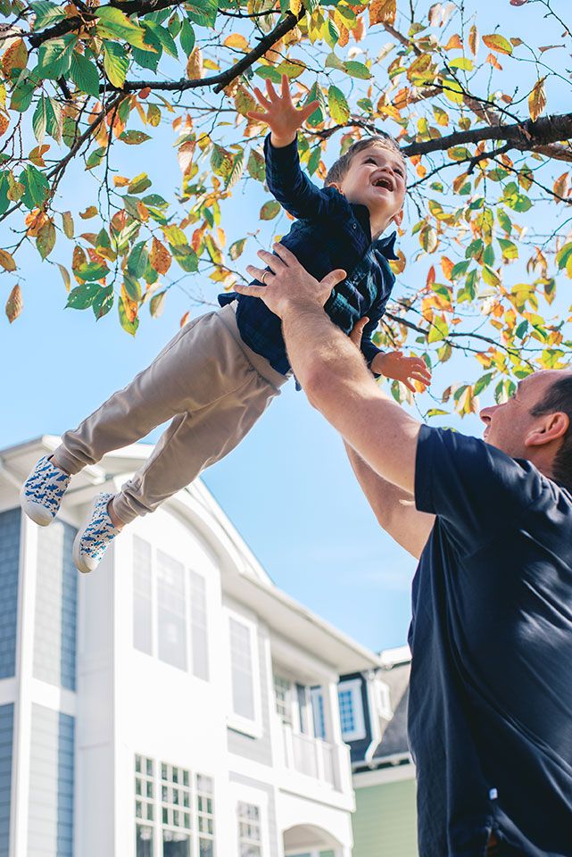 Person in navy shirt reaching up to trim tree branches in front of white house on sunny autumn day.