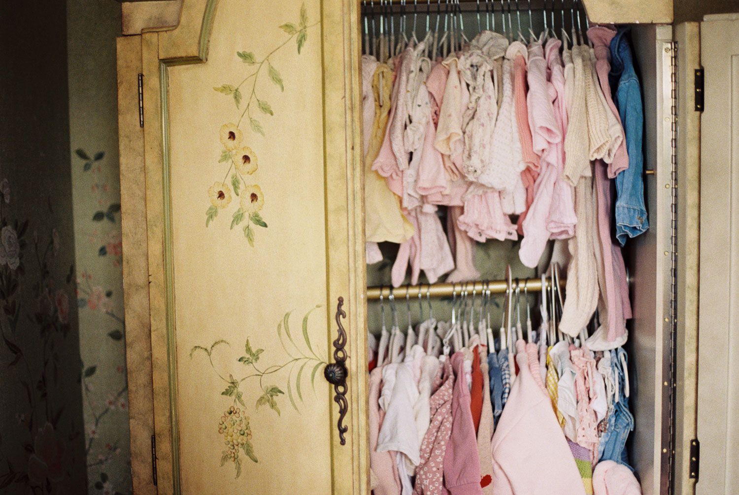 Vintage painted wardrobe open to reveal rows of pink baby girl clothes hanging neatly on hangers.