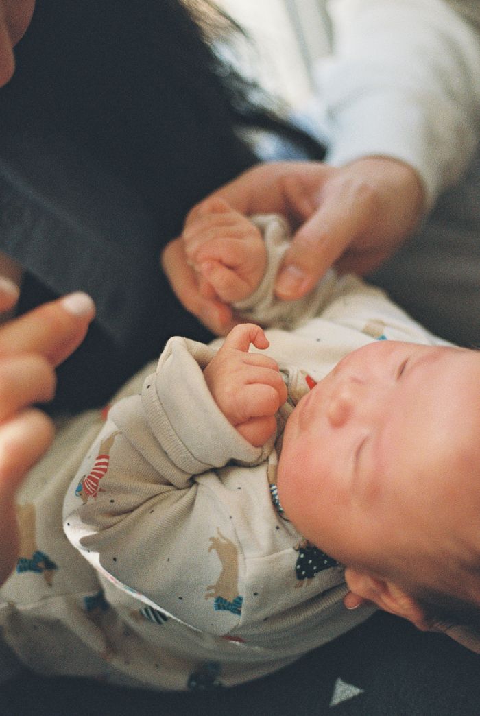A newborn baby in a printed onesie sleeps peacefully while being held by adult hands.