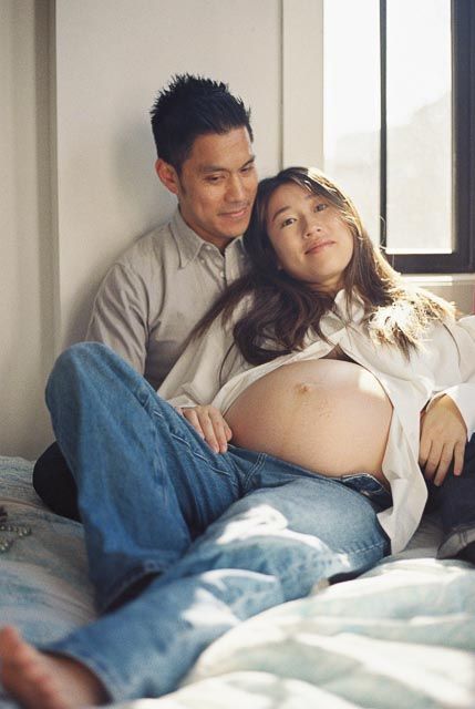 Couple sharing a tender moment, pregnant woman resting on partner's lap by a sunlit window on a bed.
