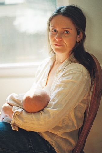 A mother sits in a wooden chair by a window, cradling her newborn baby while breastfeeding.