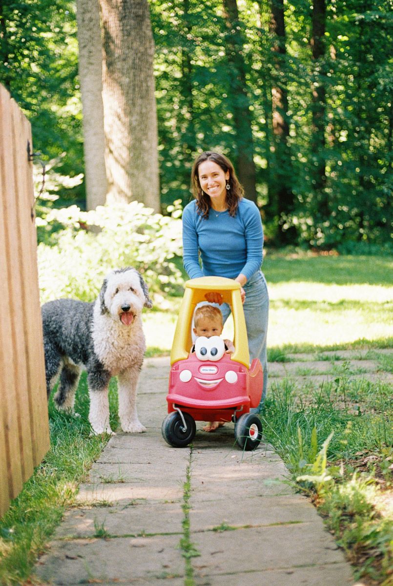 Woman smiling while pushing toddler in red Little Tikes Cozy Coupe toy car on garden path with large fluffy dog.