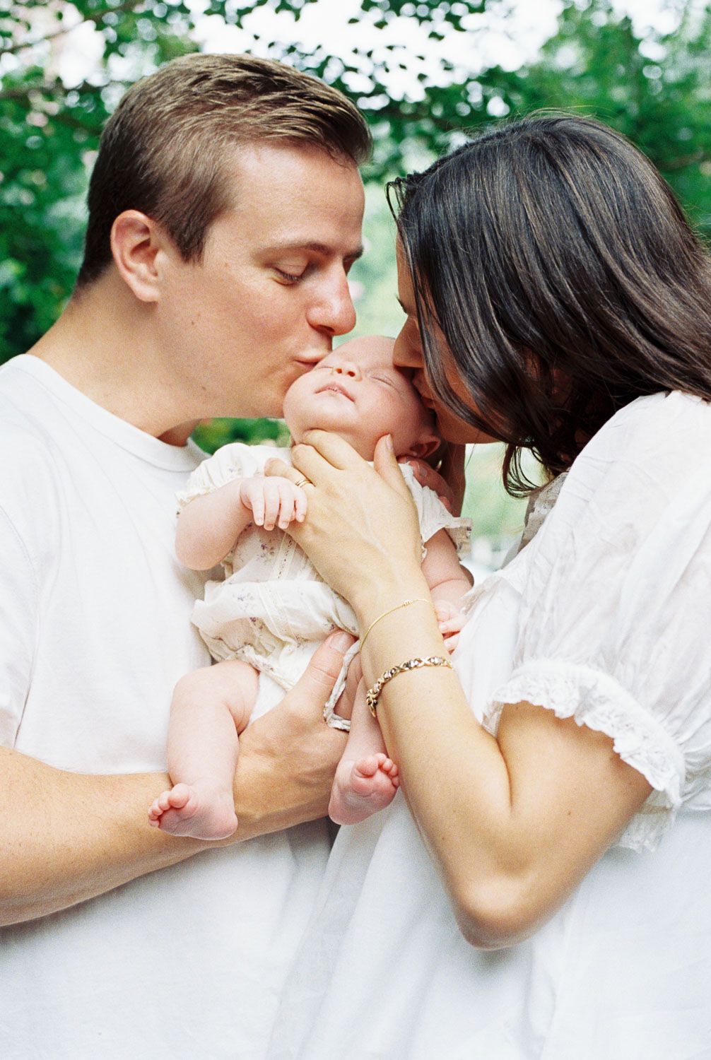 Parents in white clothing kissing their newborn baby outdoors with green trees in the background.