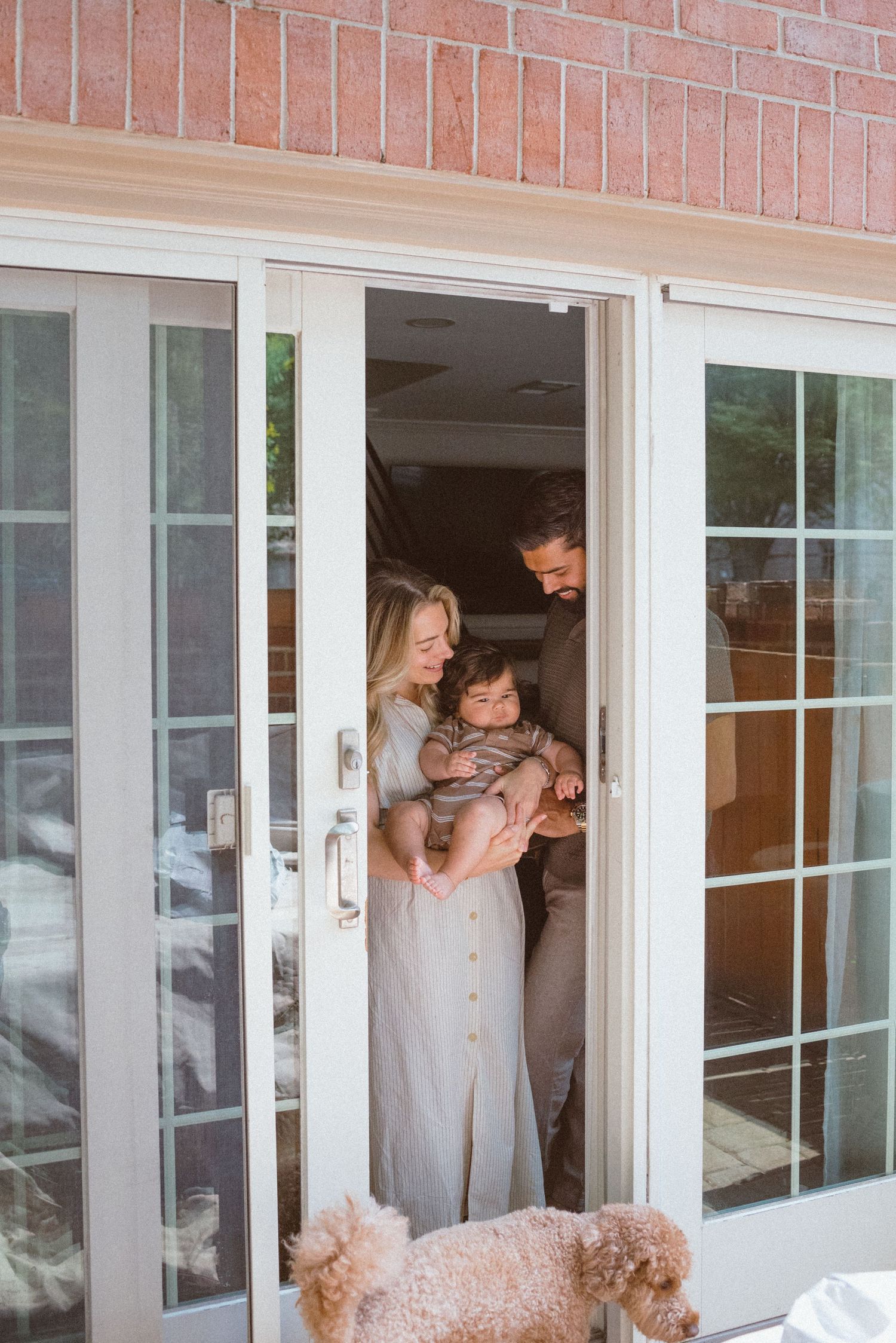 Family standing in doorway of brick home with French doors while fluffy dog waits outside on patio.