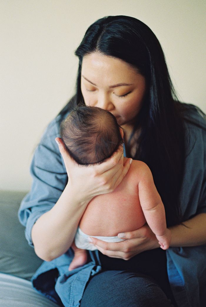 A mother with long black hair gently kisses her newborn baby on the head while cradling them in her arms.