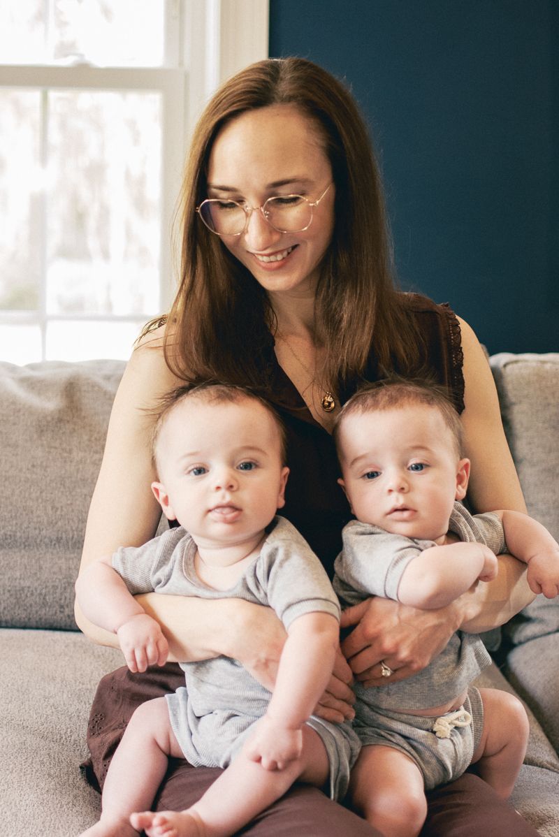 A smiling mother with glasses holds her twin babies on a gray couch in a cozy home setting.