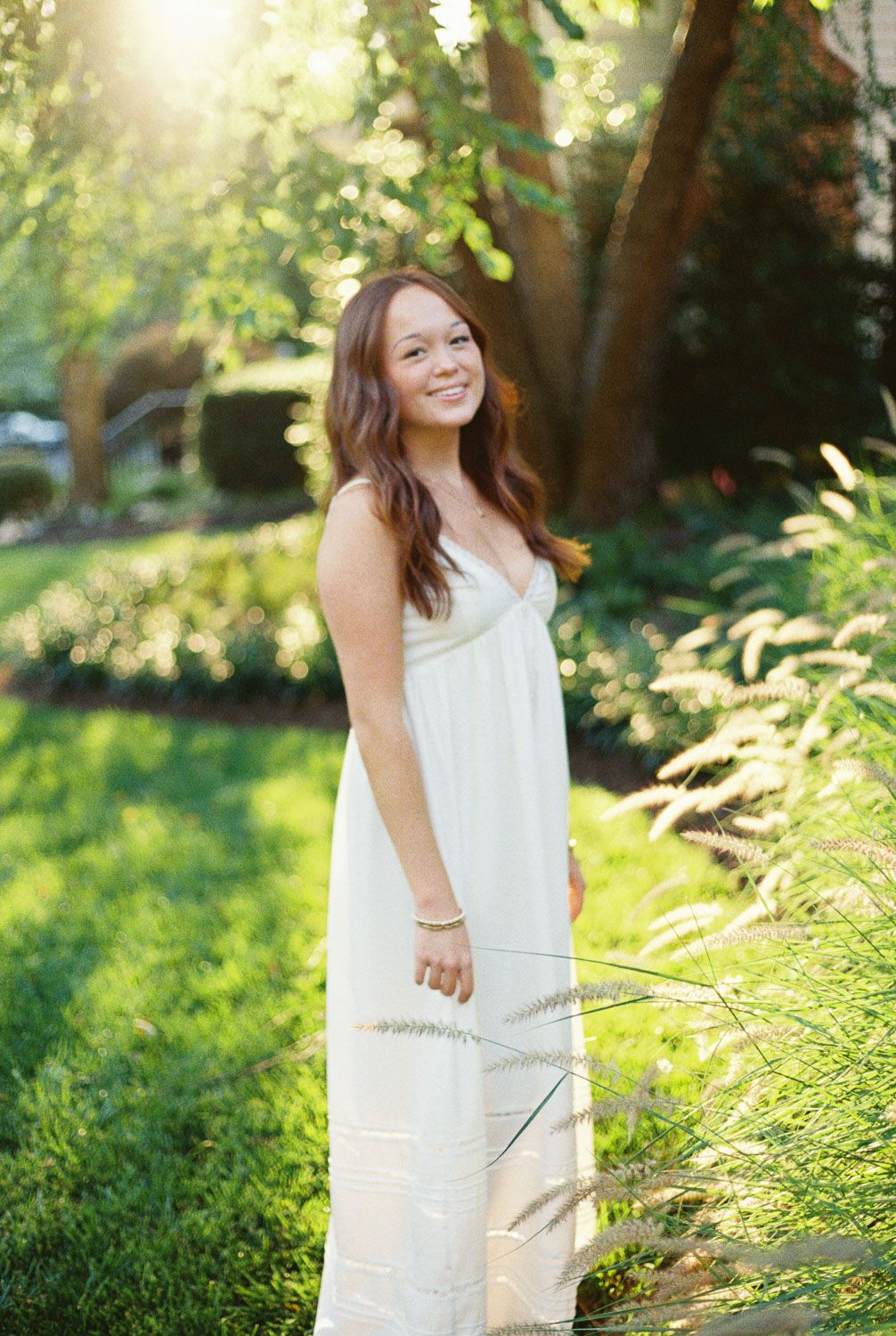 Smiling young woman with long wavy brown hair in a white sundress stands in a sunlit garden beside tall ornamental grasses.