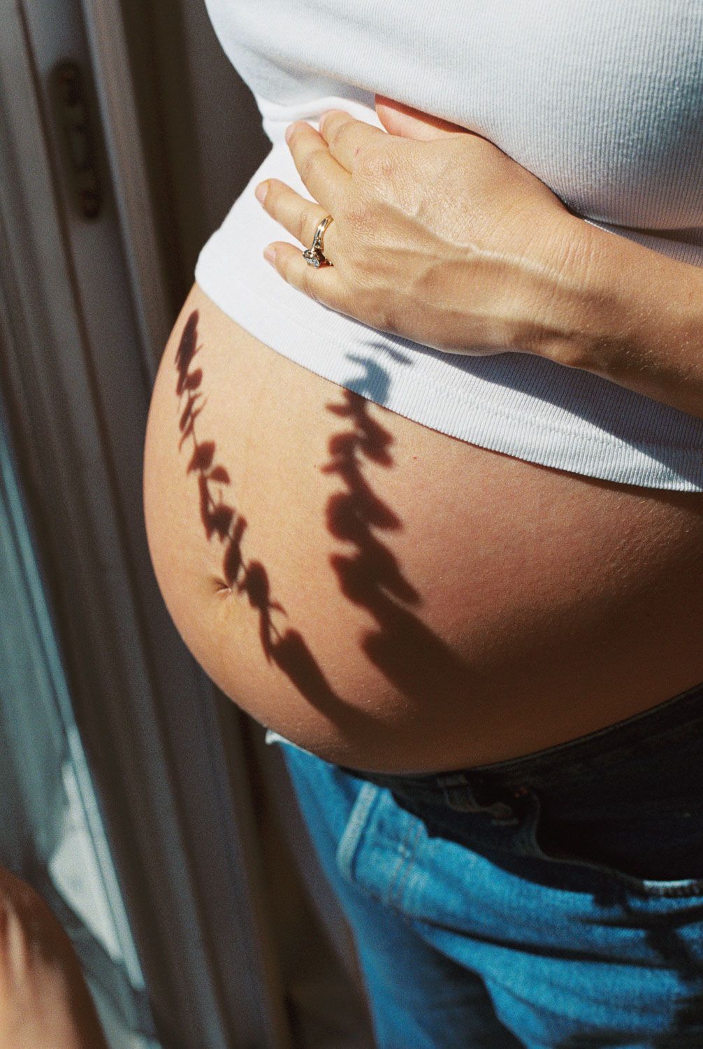 Close-up of a pregnant woman's bare baby bump in sunlight, wearing a white crop top and blue jeans.