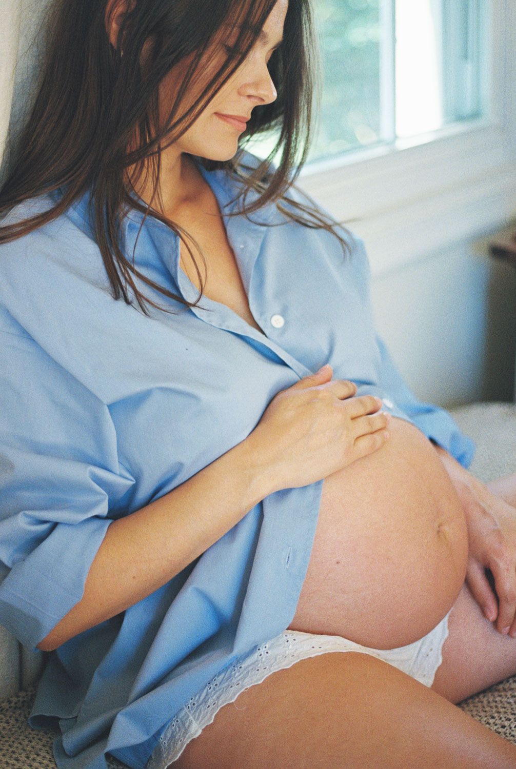 Pregnant woman in blue shirt tenderly cradling her baby bump while sitting by a bright window.