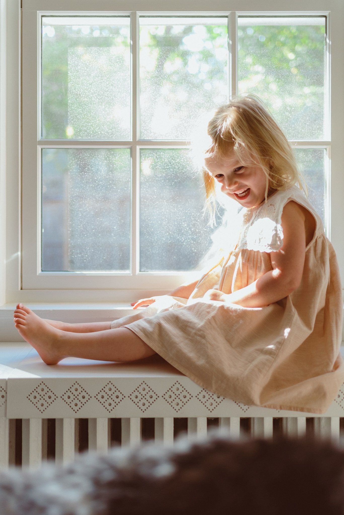 A child in a white dress sits on a window sill with sunlight streaming through paned glass windows.