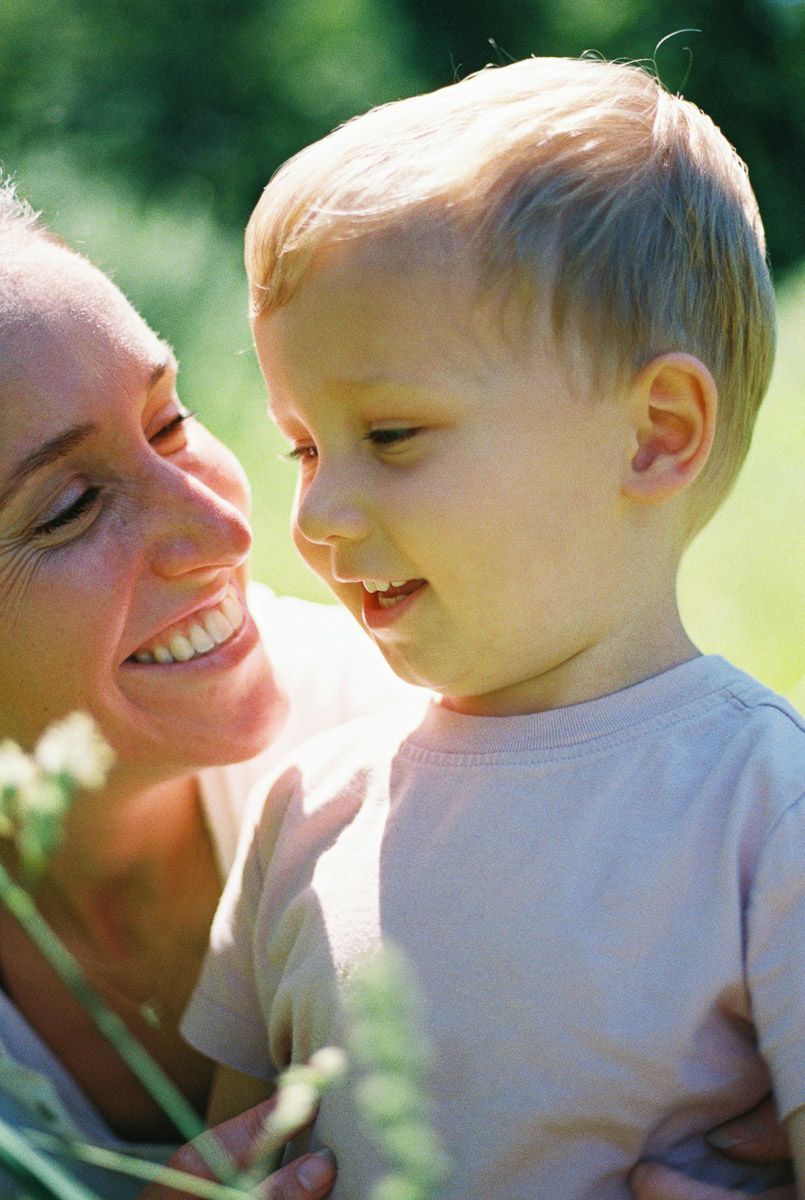 A smiling woman holds a happy blonde toddler boy outdoors in a sunny green garden setting.