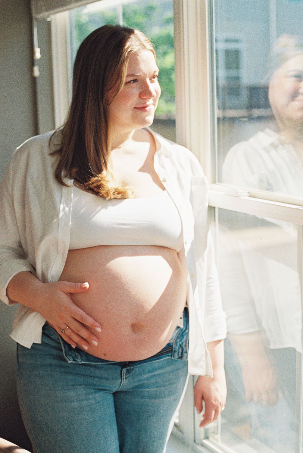 Pregnant woman in white shirt and jeans cradling baby bump while standing by a sunlit window indoors.