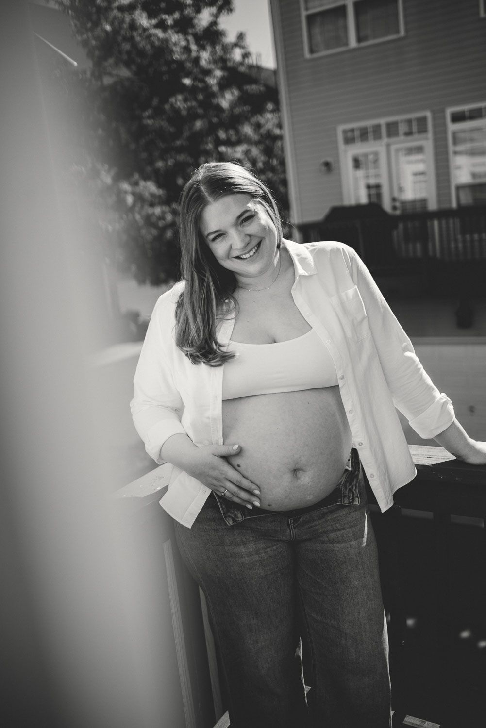 Smiling pregnant woman in white shirt and jeans holds baby bump outdoors in black and white maternity photo.