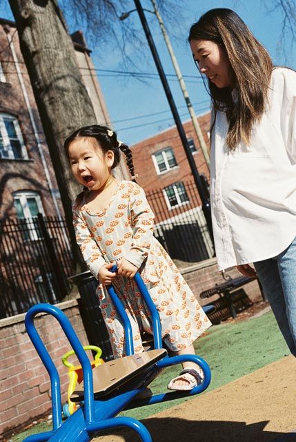 Young girl laughing on playground seesaw while mother watches on sunny day.