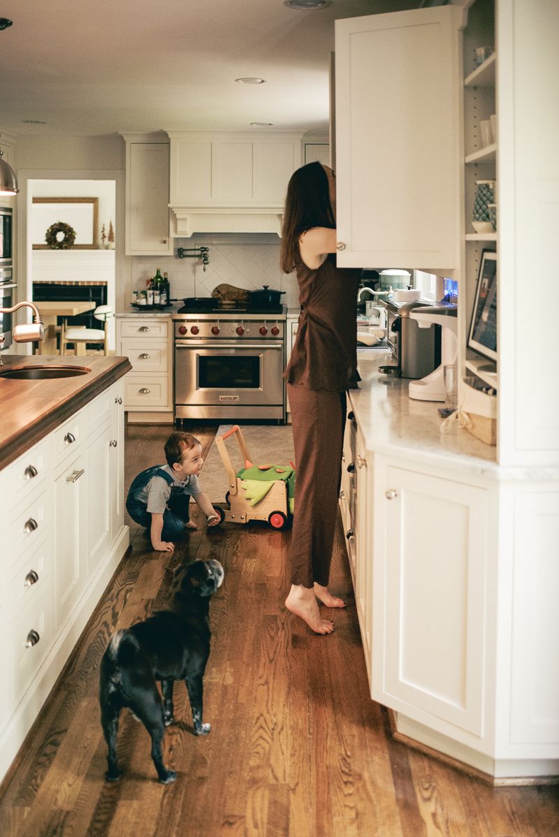 A woman stands barefoot in a white kitchen while a young child plays on hardwood floors beside a black Labrador dog.