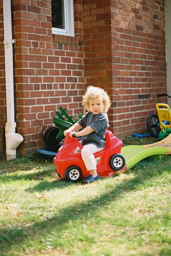 Curly-haired toddler sitting on a red ride-on toy car in a sunny backyard next to a brick house.