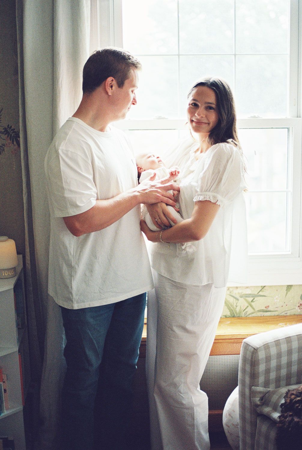New parents in white clothing stand by a bright window, lovingly holding their newborn baby in a cozy nursery room.