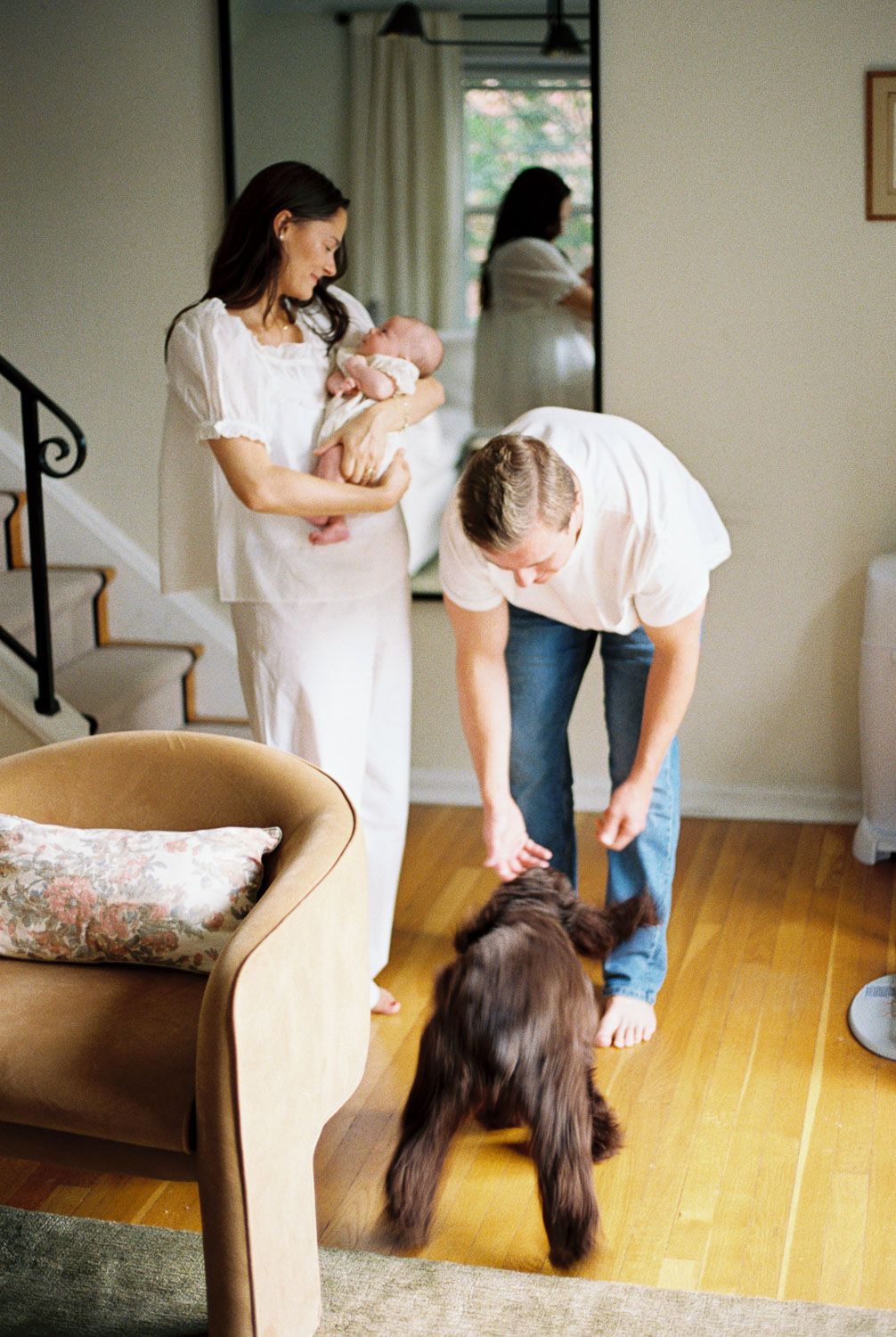 A young family with a newborn baby and their brown dog gather in a bright, modern living room with hardwood floors.