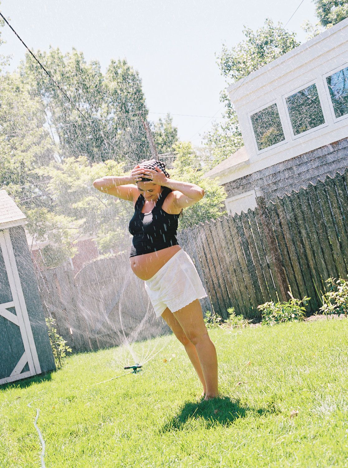 Pregnant woman in black top and white skirt cooling off in backyard sprinkler on sunny summer day.